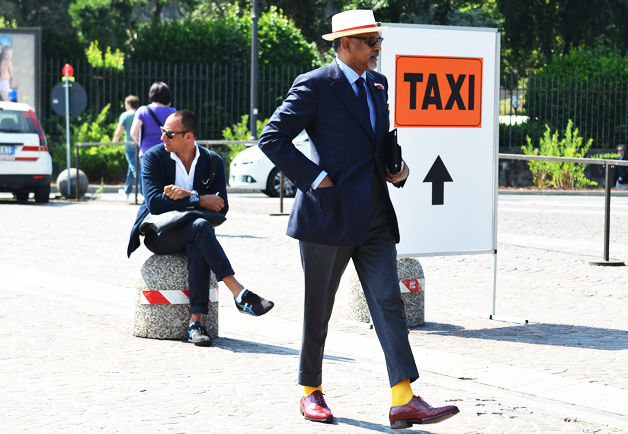 homme portant une veste de costume bleu marine un pantalon gris des chaussettes jaunes des chaussures marron et un chapeau blanc et rouge