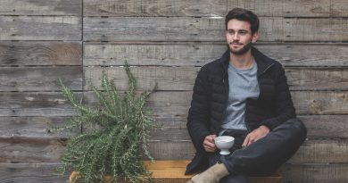 homme brun assis sur un banc portant un jean un t shirt gris une doudoune foncée et des boots beige une tasse à la main