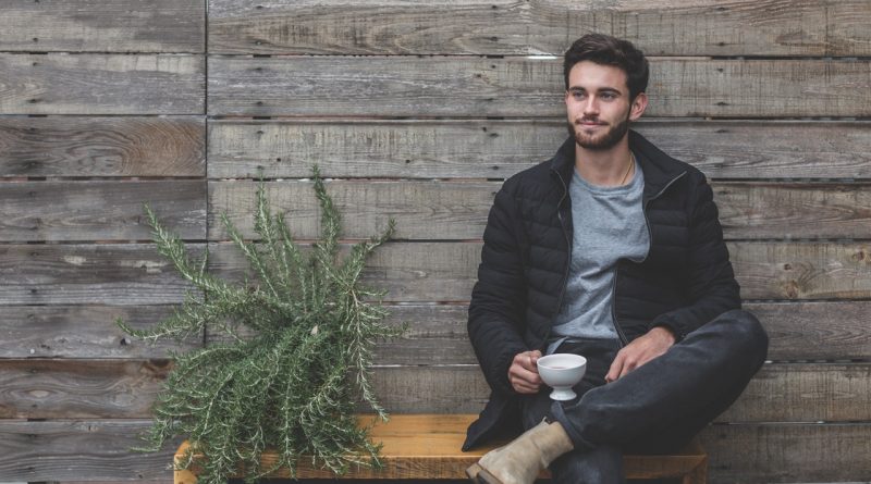 homme brun assis sur un banc portant un jean un t shirt gris une doudoune foncée et des boots beige une tasse à la main